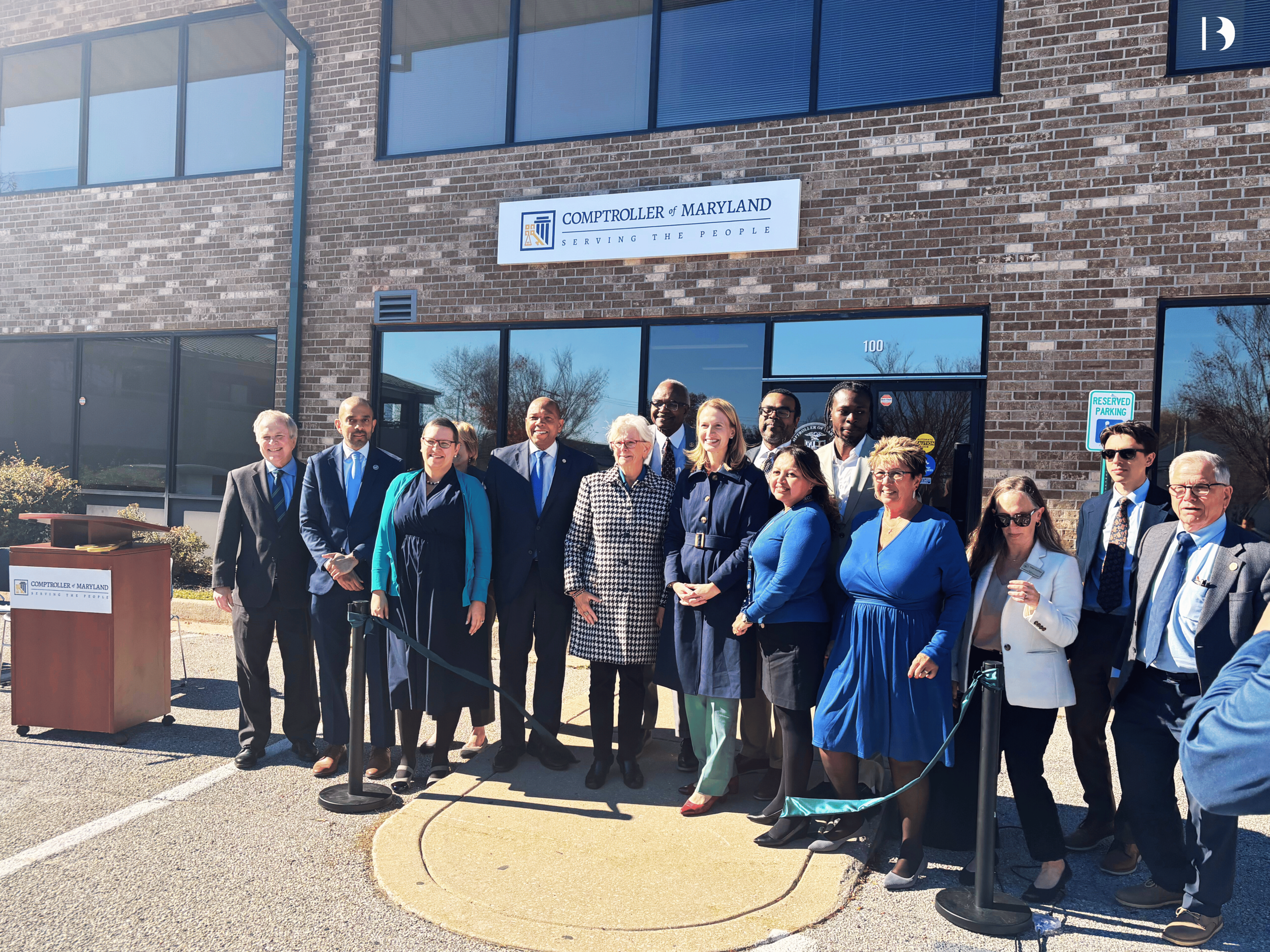 Comptroller Brooke E. Lierman stands alongside state and local officials in front of the new Maryland Comptroller’s Baltimore County Branch Office at Executive Park West in Windsor Mill. The group poses for a ribbon-cutting photo outside the brick office building, with the Comptroller of Maryland sign visible above the entrance and blue skies overhead.
