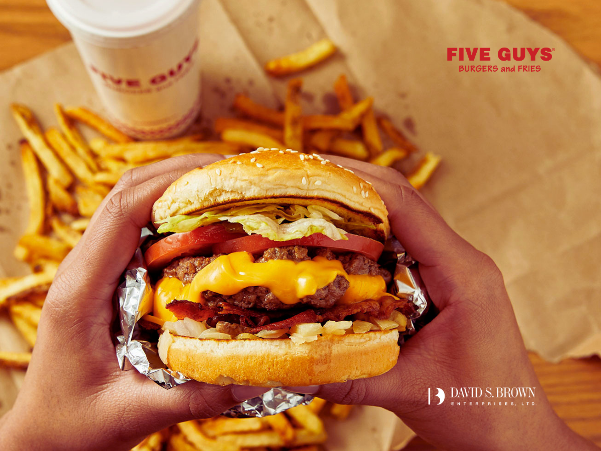 Hands holding a freshly made Five Guys cheeseburger topped with lettuce, tomato, bacon, and melted cheese, with hand-cut fries and a drink in the background. Promotional image announcing Five Guys is now open at Lockwood Place in Downtown Baltimore, featuring David S. Brown Enterprises branding.