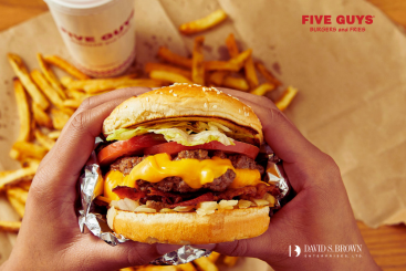 Hands holding a freshly made Five Guys cheeseburger topped with lettuce, tomato, bacon, and melted cheese, with hand-cut fries and a drink in the background. Promotional image announcing Five Guys is now open at Lockwood Place in Downtown Baltimore, featuring David S. Brown Enterprises branding.