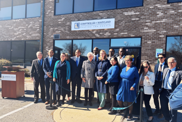 Comptroller Brooke E. Lierman stands alongside state and local officials in front of the new Maryland Comptroller’s Baltimore County Branch Office at Executive Park West in Windsor Mill. The group poses for a ribbon-cutting photo outside the brick office building, with the Comptroller of Maryland sign visible above the entrance and blue skies overhead.